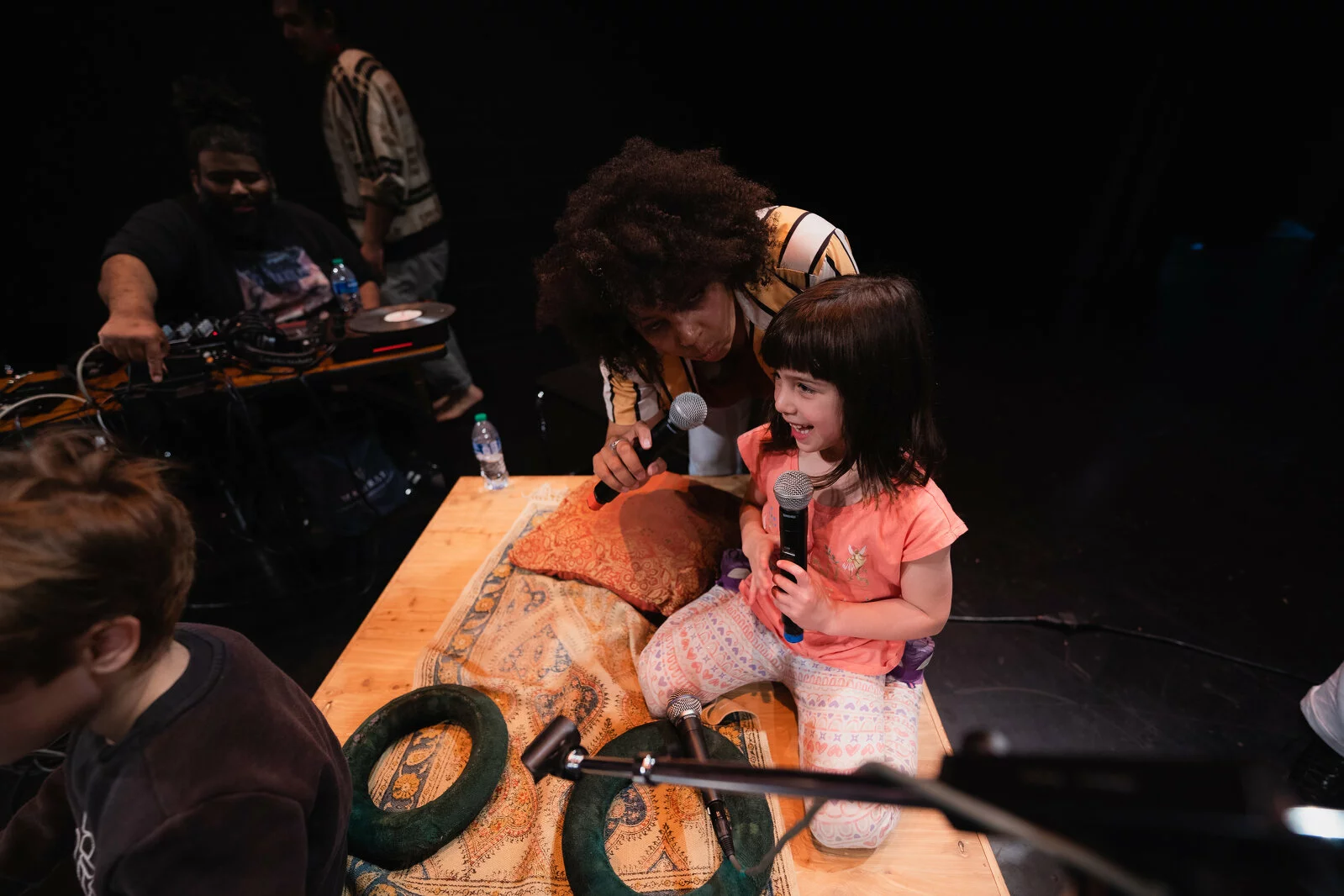A child and an adult lean in together with microphones in their hands. Behind them a man sits at a table with a record player and sound equipment.