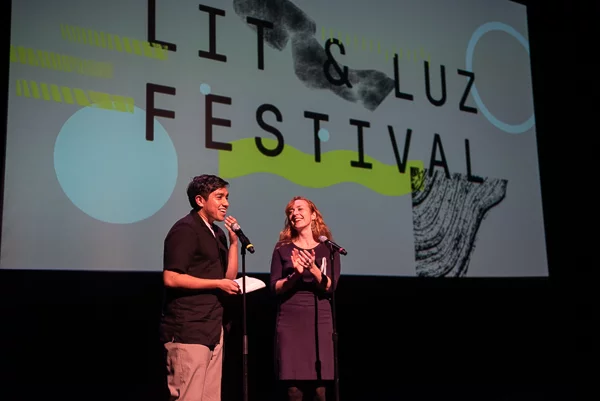 Two speakers with microphones on a Stage with a LIT & LUZ FESTIVAL screen behind them.