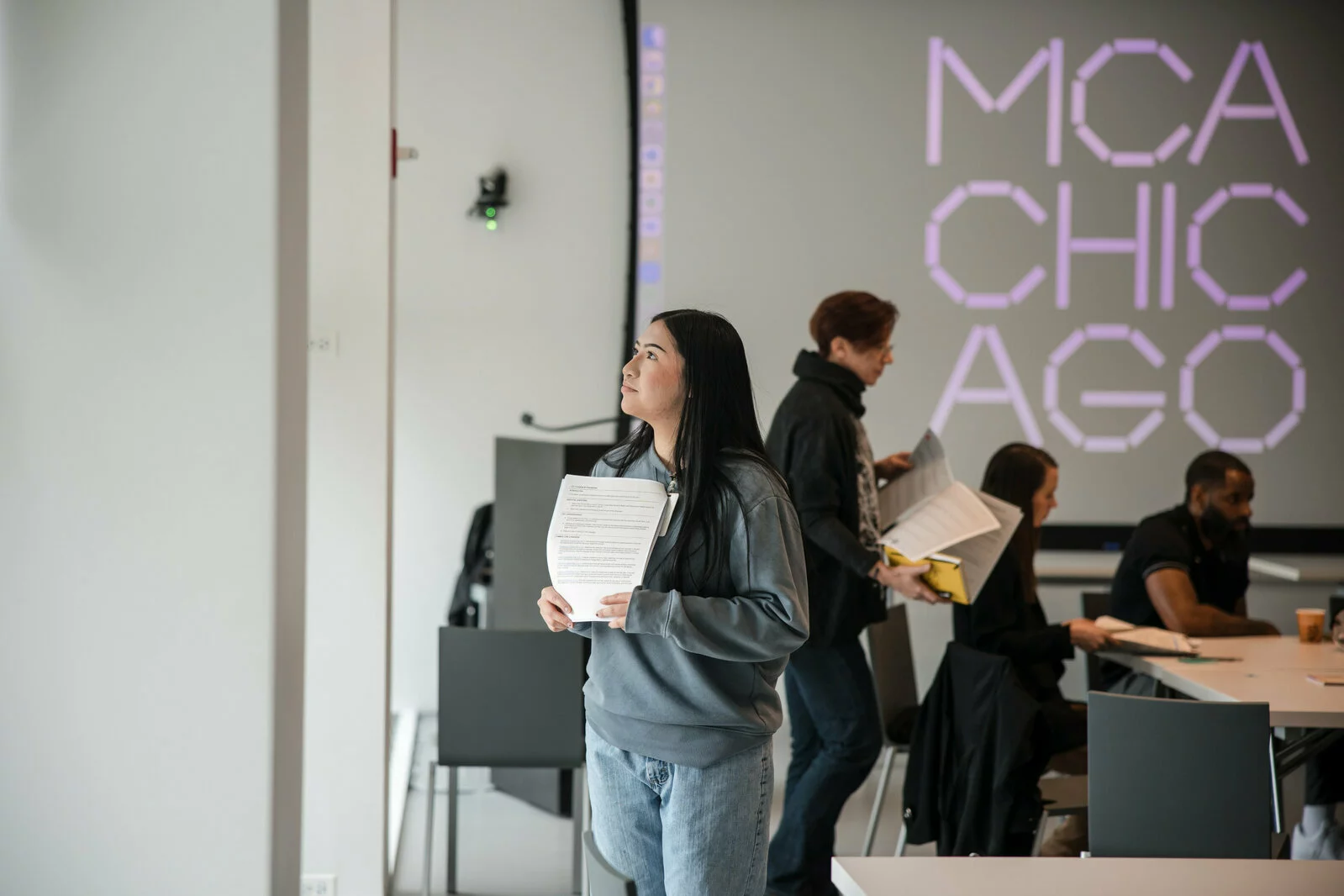 A woman stares up at something off camera while behind her a group of people gather at tables in front of a big screen displaying the MCA Chicago logo.