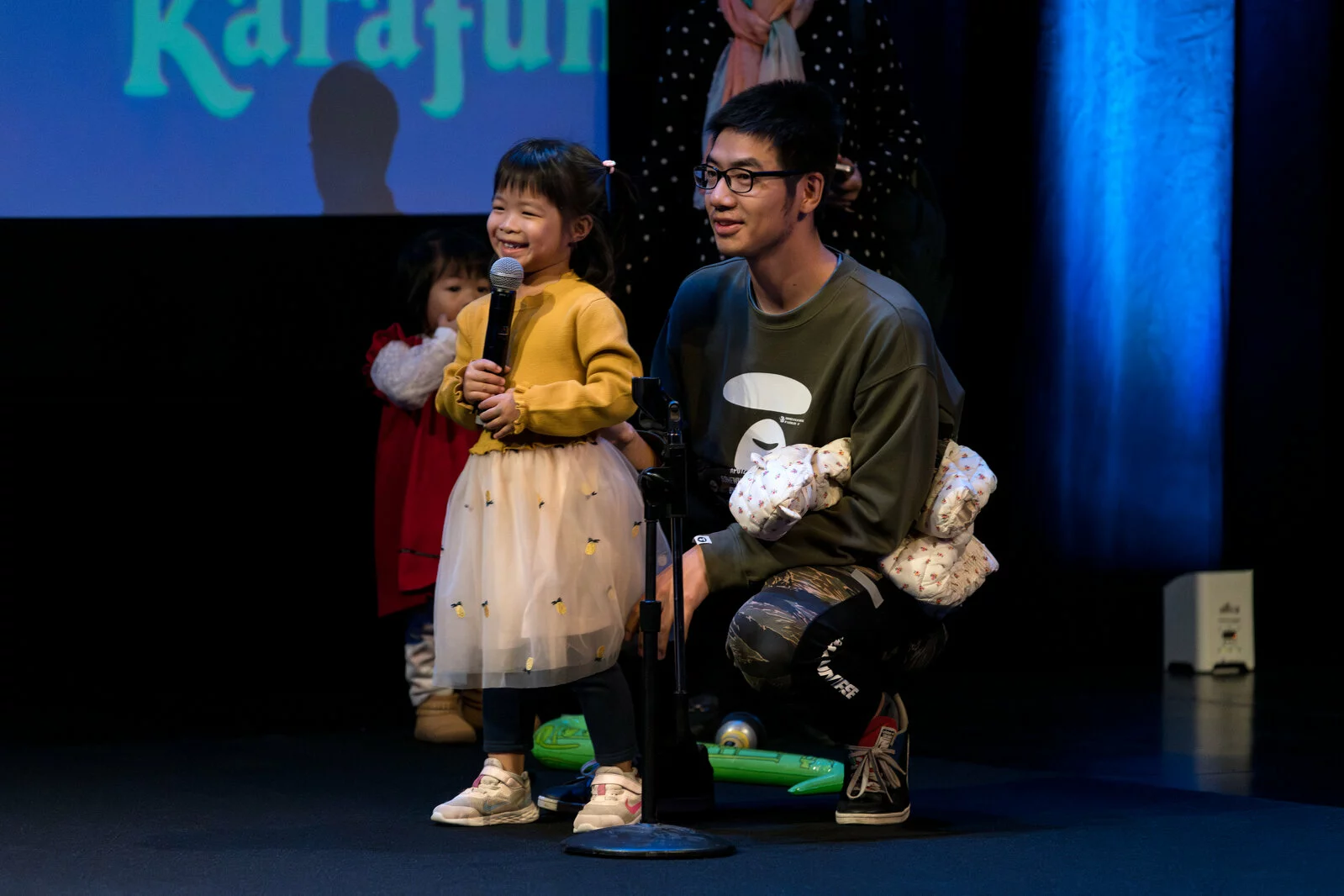 A child smiles while holding a microphone as their adult crouches beside them on a darkened stage.