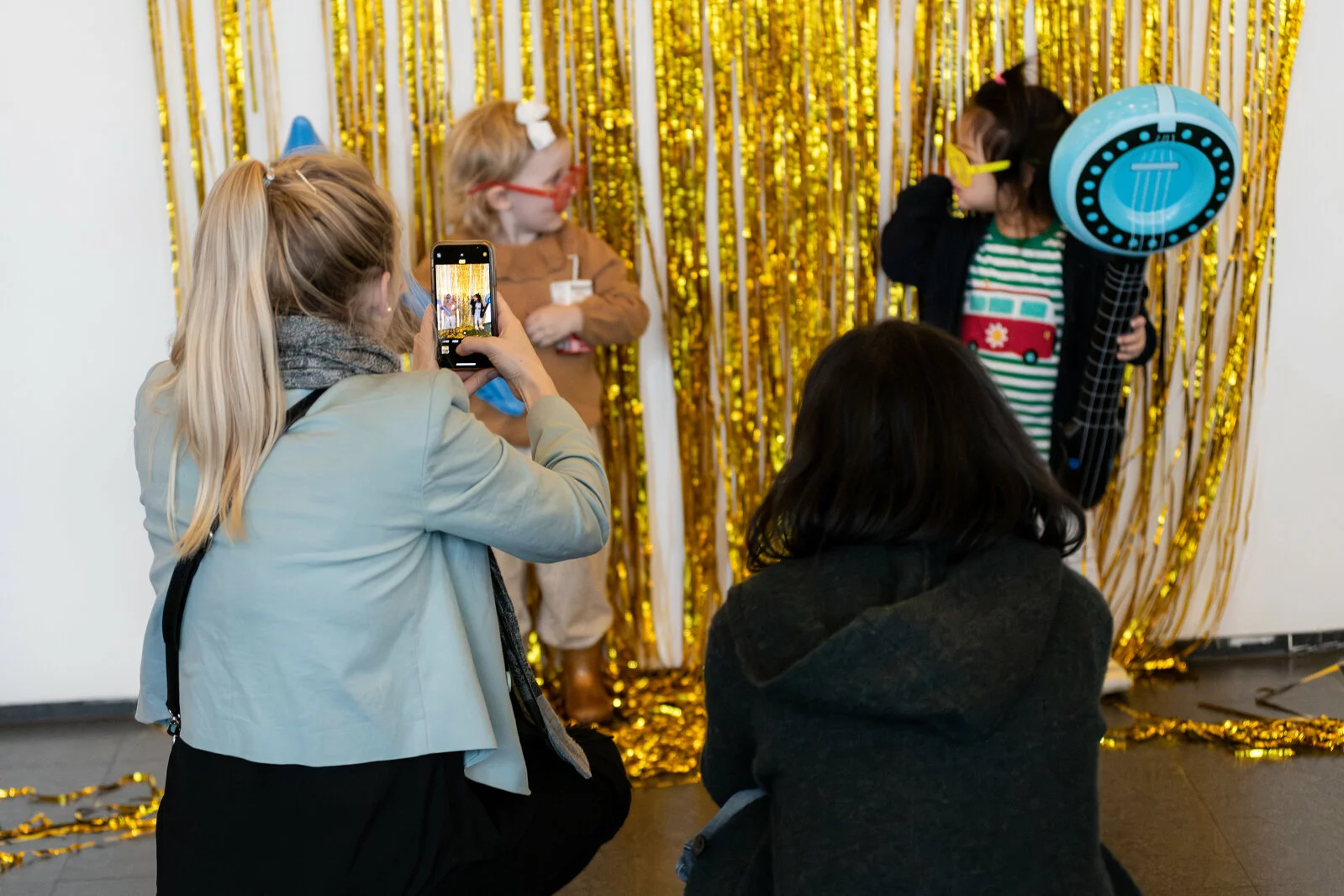 Two children pose in front of gold streamers as their adults take photos of them.