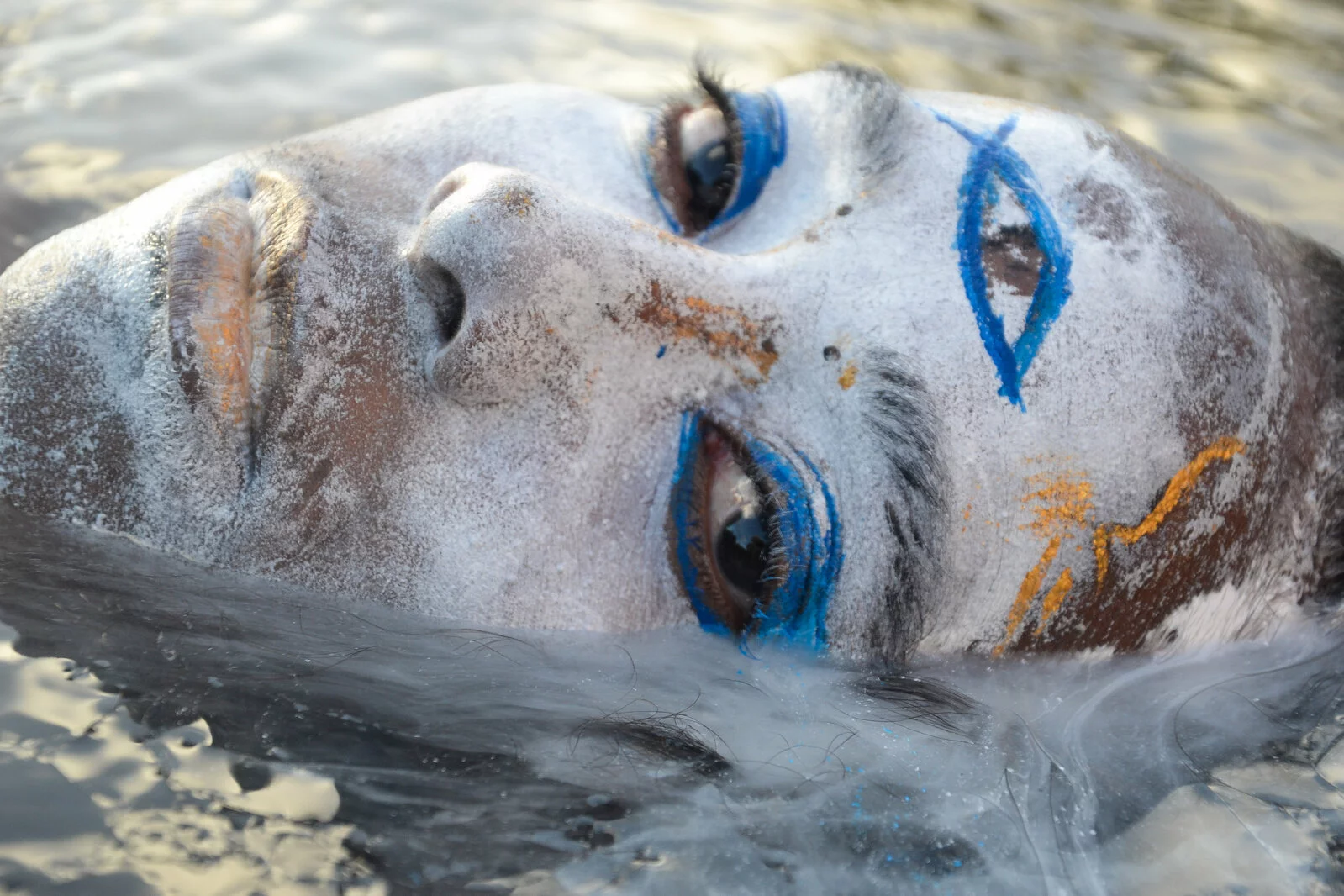 Close up of a person's head in a reclined position turned slightly towards the camera. Their face is painted white with blue eyeshadow and a painted third eye.