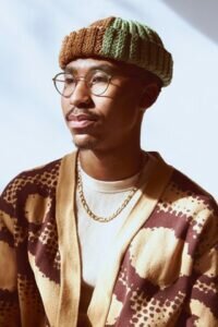 Portrait of a Black man wearing glasses and a multicolored beanie against a white wall.