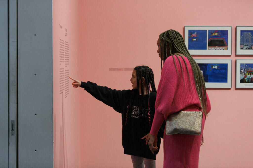 A younger person points to text on a pink wall and the guardian they are with leans in to read.