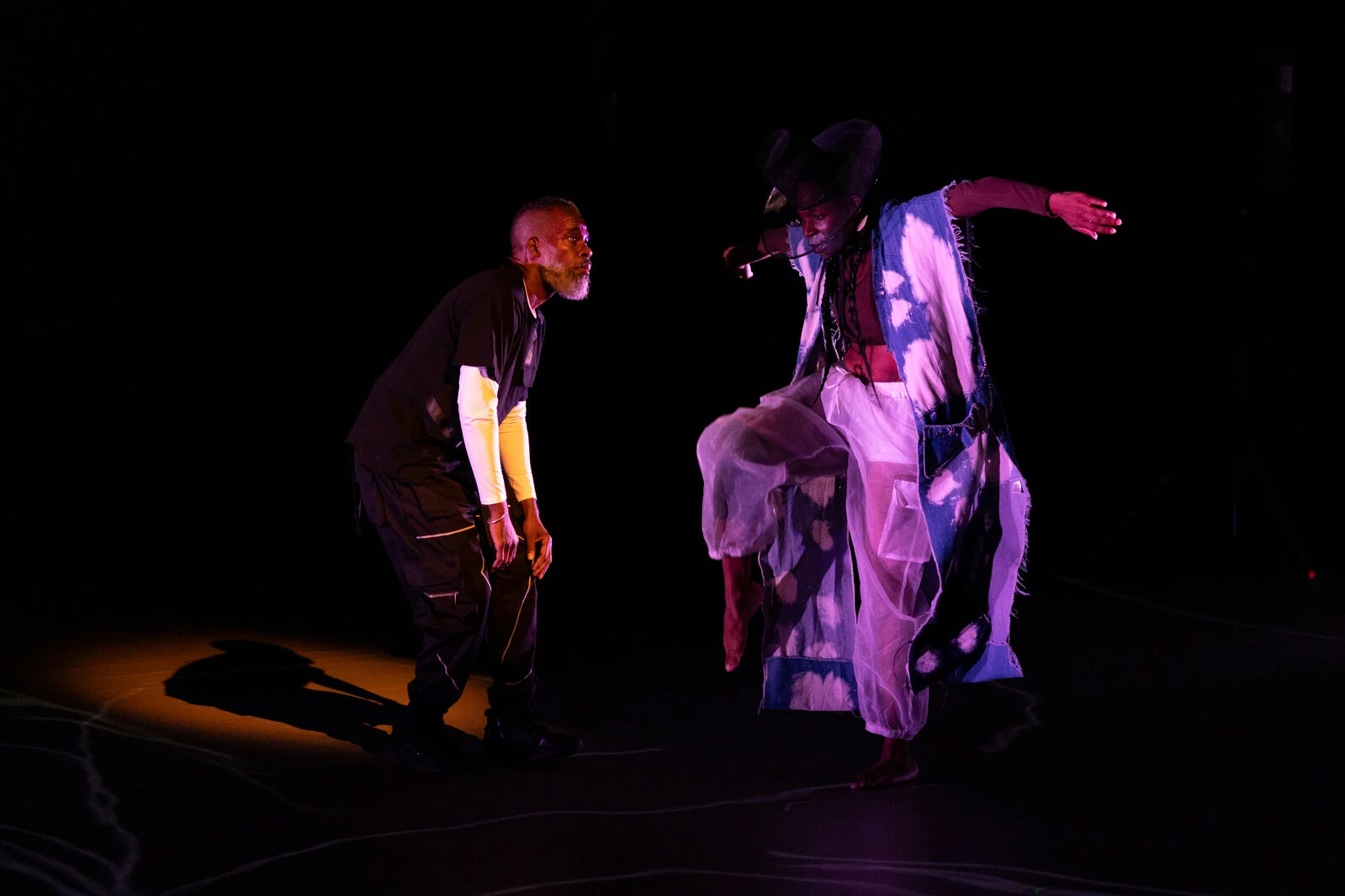 On a darkened stage lit only by purple spotlights a woman is captured mid-jump as a man crouches next to her.