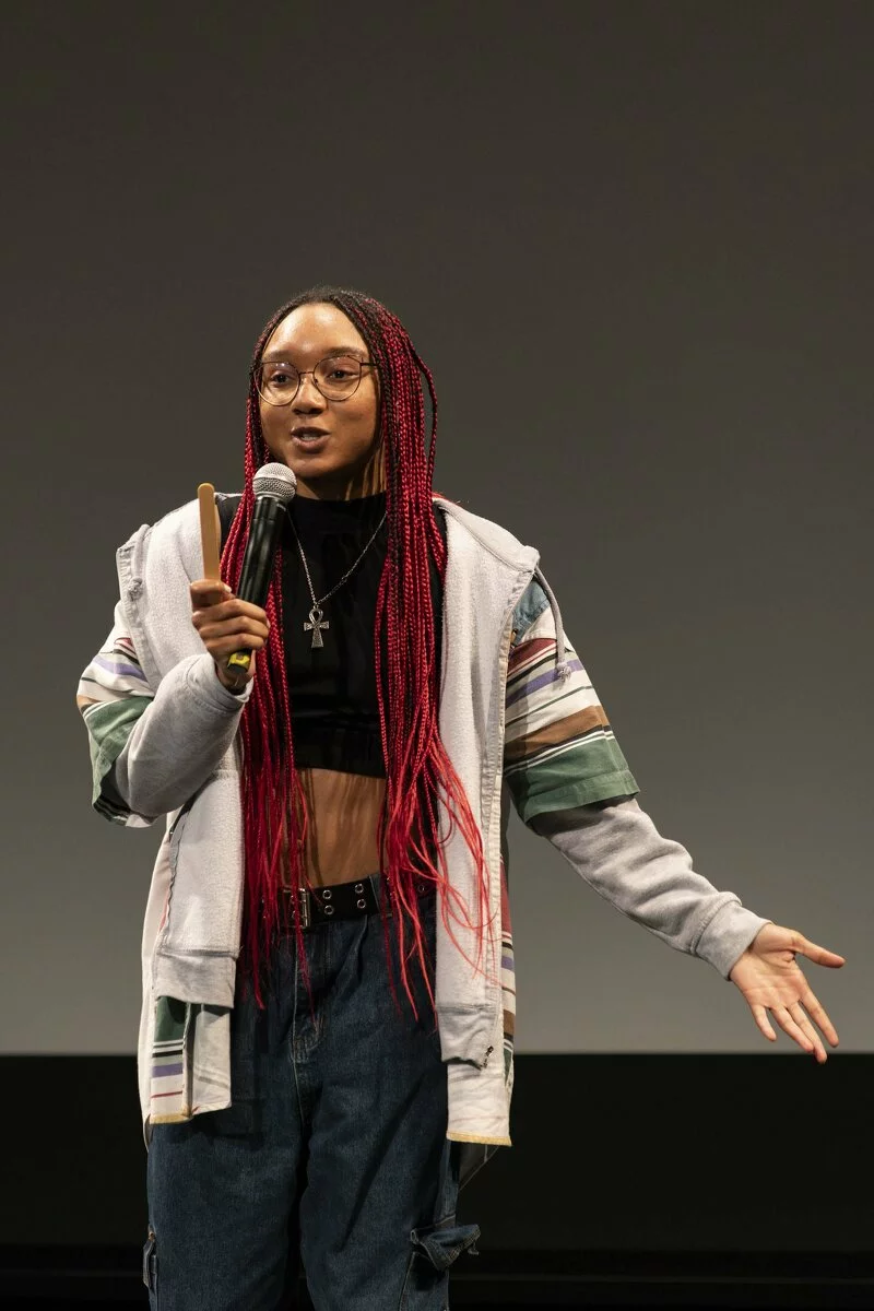 A young Black performer brandishes their left hand while holding a microphone to their mouth with the right hand.