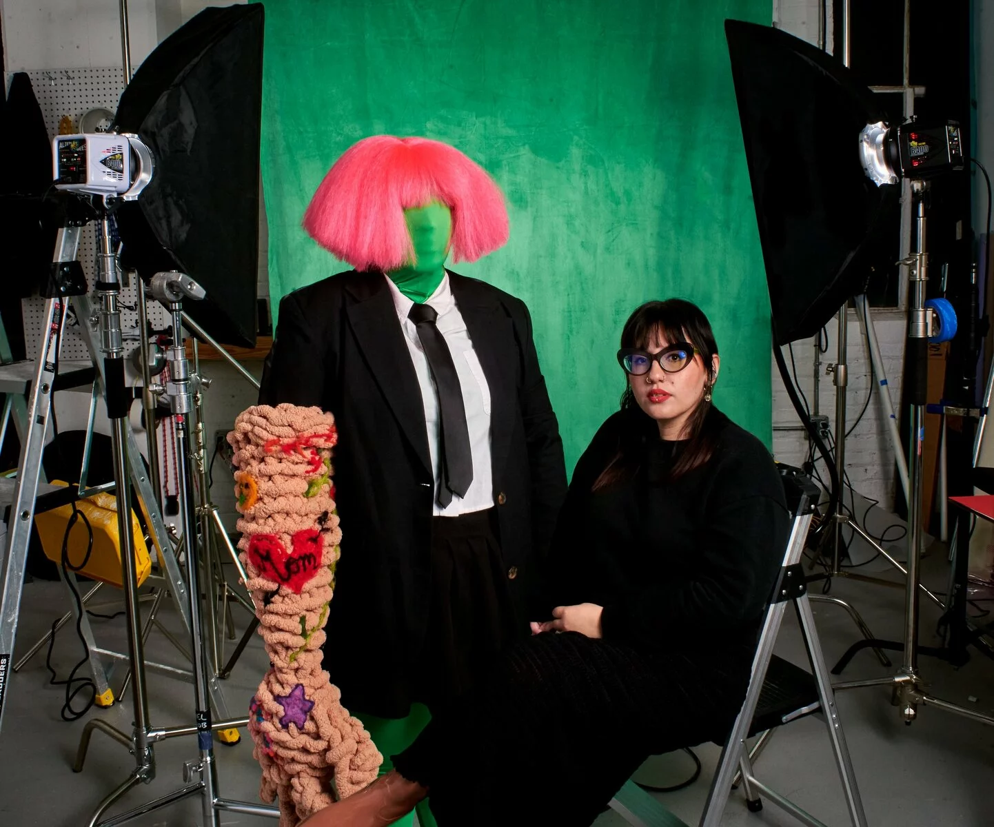 Ariella Granados sits in front of a green screen in a photography studio surrounded by props and equipment.