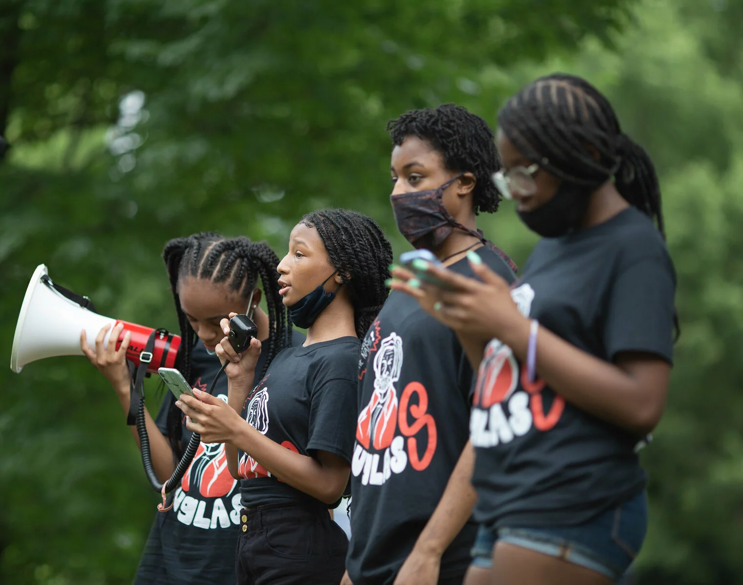 A row of four young Black adults wearing the same t-shirt. One person holds a megaphone while another speaks into its attached microphone.
