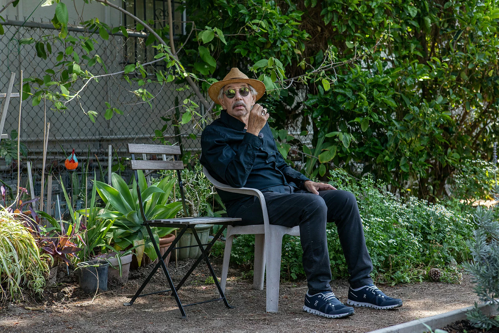 Artist David Lamelas sits on a plastic chair surrounded by green foliage.