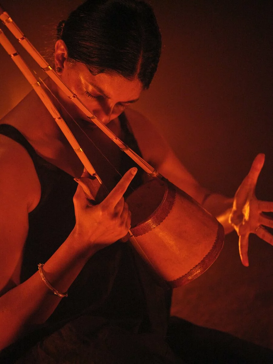 Artist Samita Sinha playing the ektara in an amber-red light, with all fingers of her left hand fully extended.