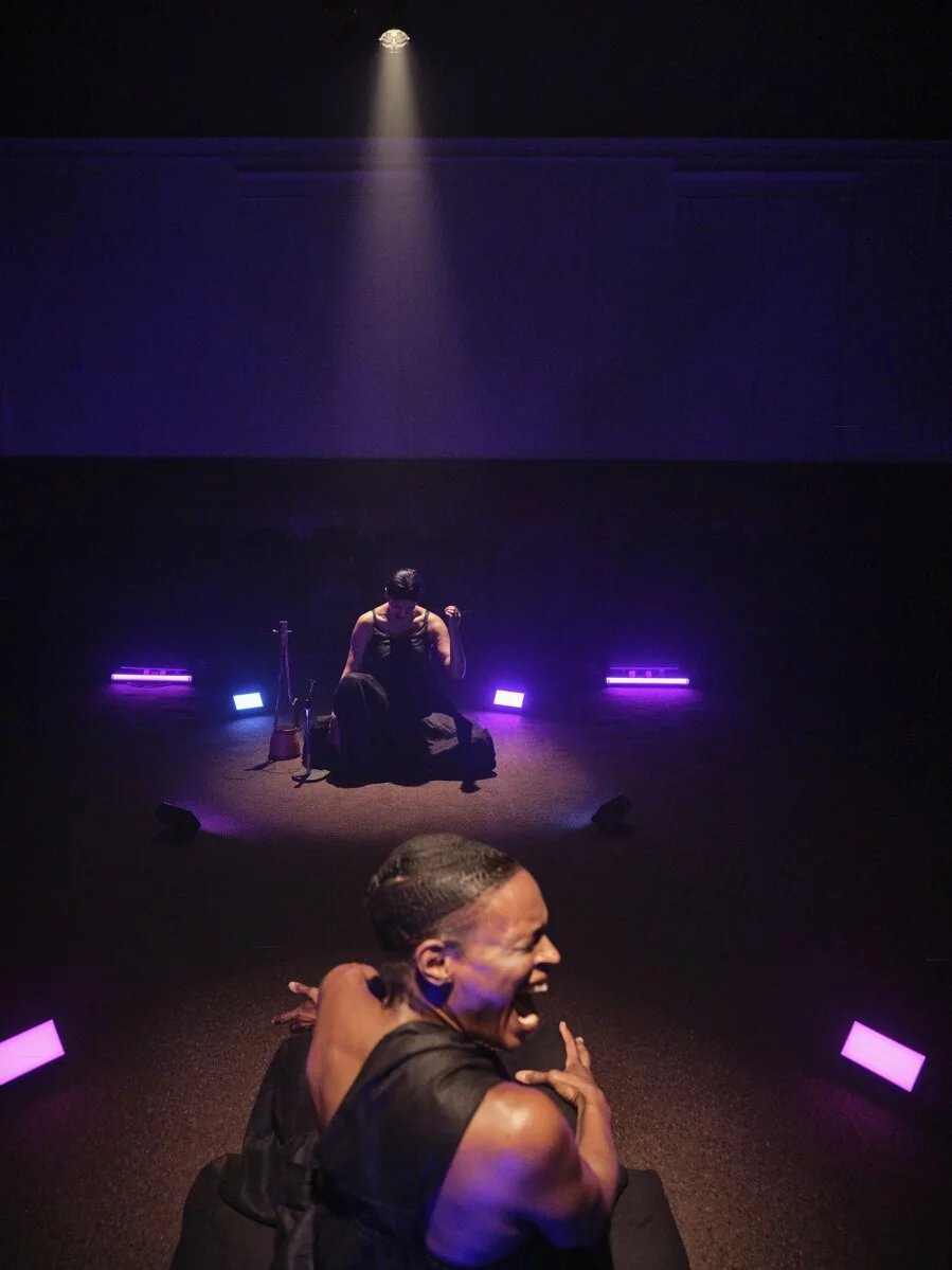 Okpokwasili (foreground) and Sinha (background) are seated across a room from each other in violet light, vocalizing together.
