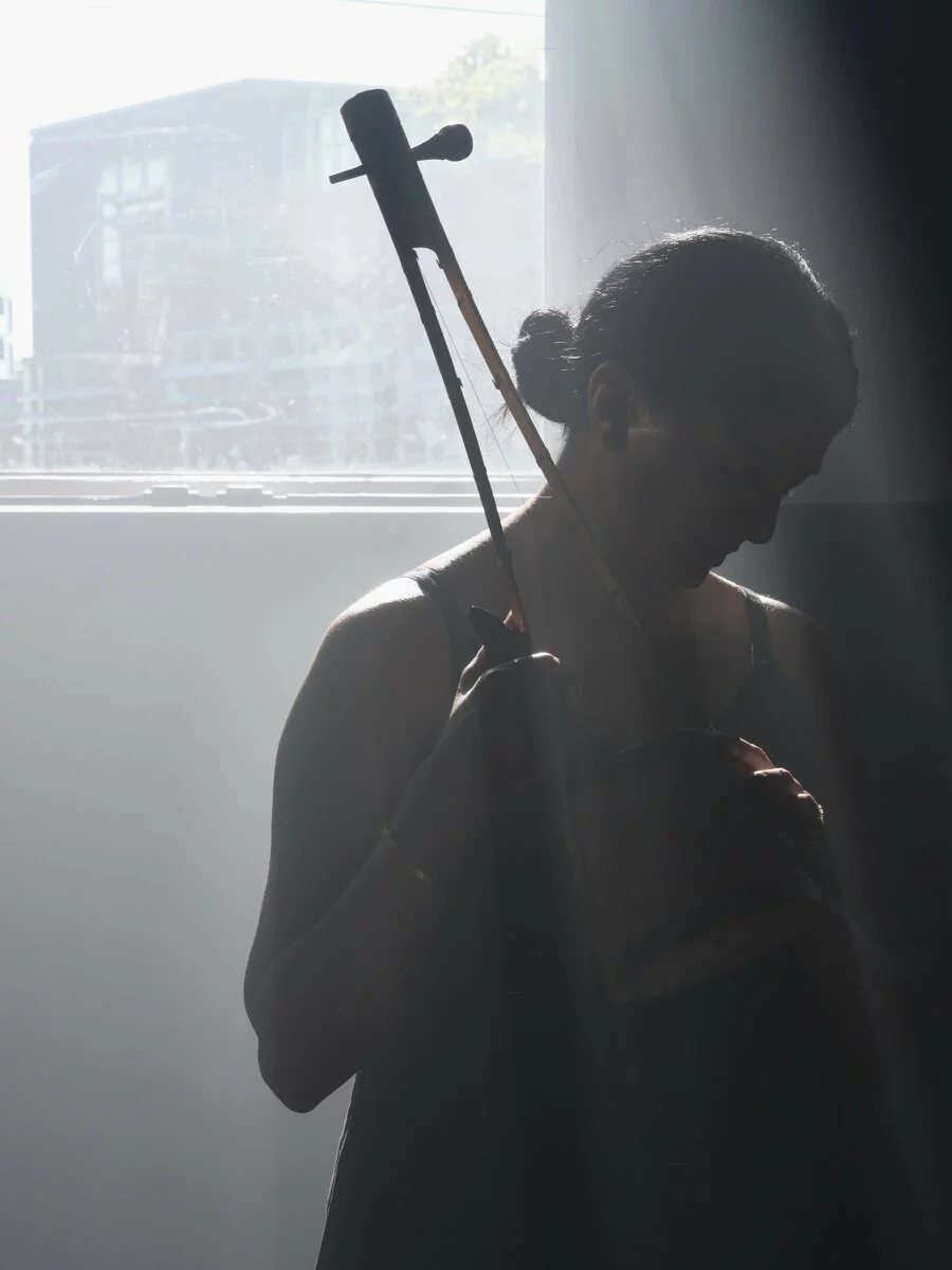 Artist Samita Sinha stands backlit, in silhouette, in front of a window, holding an ektaara, a one-stringed instrument.
