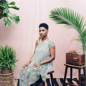 Portrait of a Black woman sitting in a pink room filed with plants and an old radio.