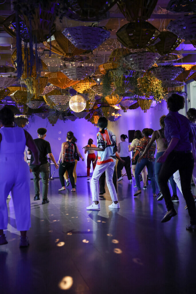 A group line dances under a disco ball in a blue-lit room with plants hanging from the ceiling.
