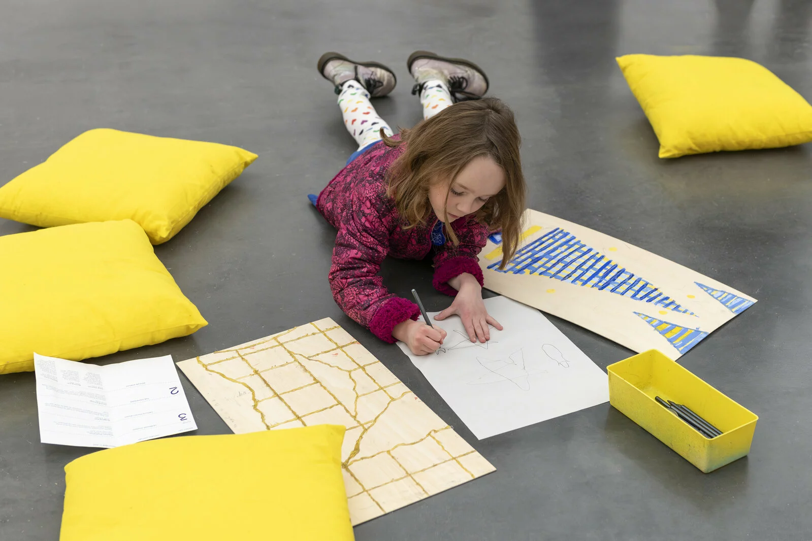 A child lays on a concrete floor and colors while surrounded by yellow pillows and sheets of paper