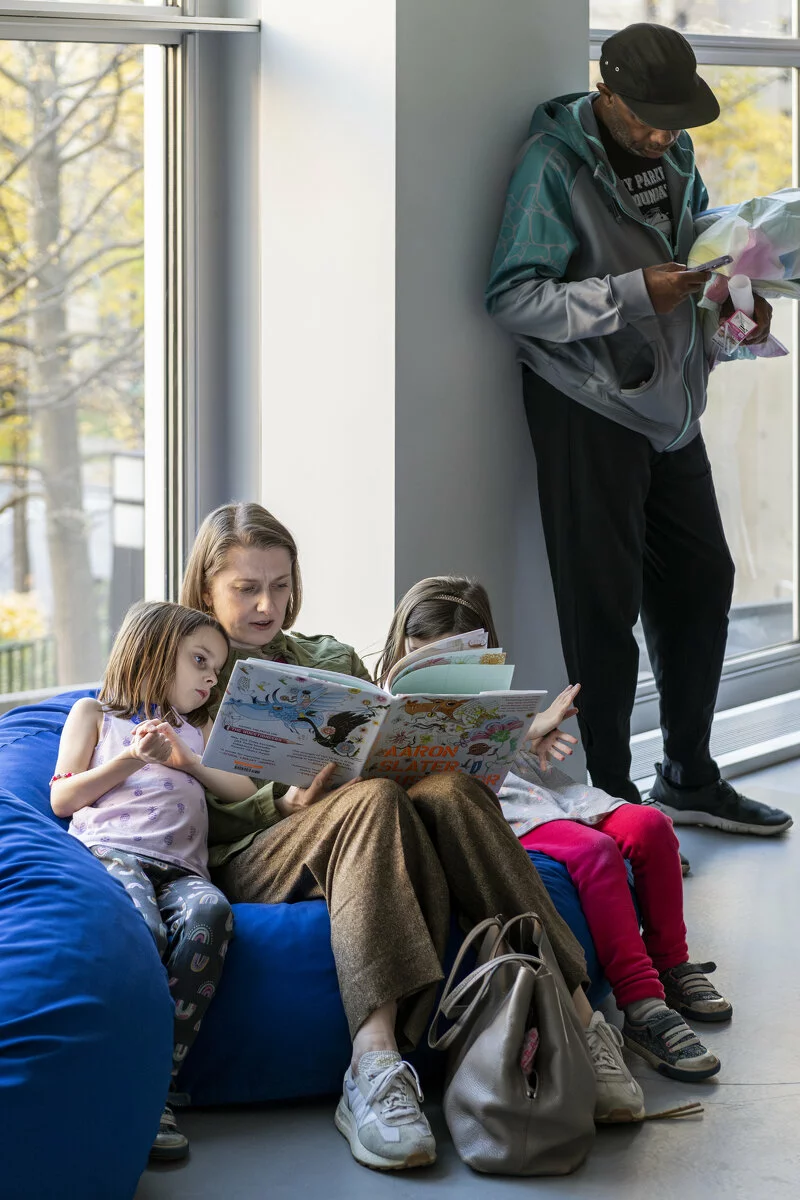 An adult and two children sit on blue bean bags together reading a book while another adult stands reading against a wall.