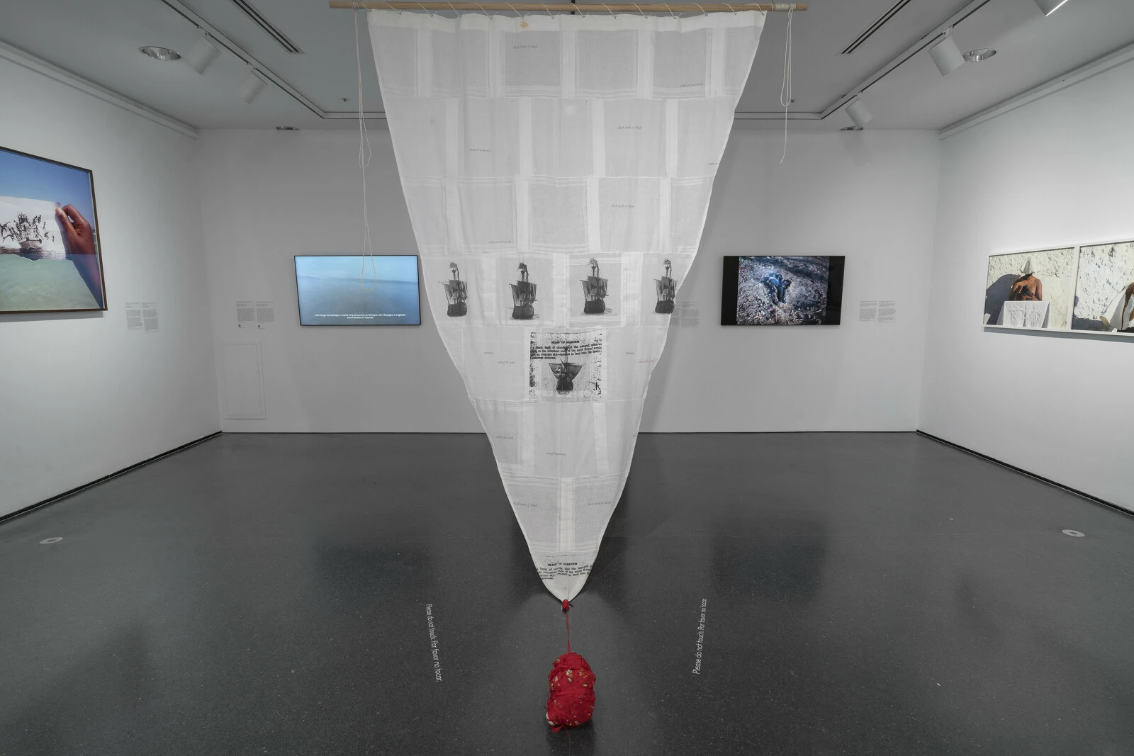 Gallery with several photographs, videos, and other artworks on the wall. In the center a sail-like object hangs from the ceiling.