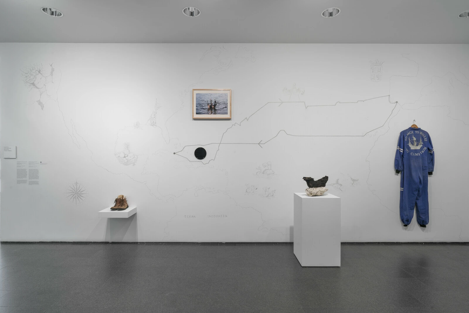 One wall of a gallery space filled with a map and objects, including shoes and a jumpsuit.