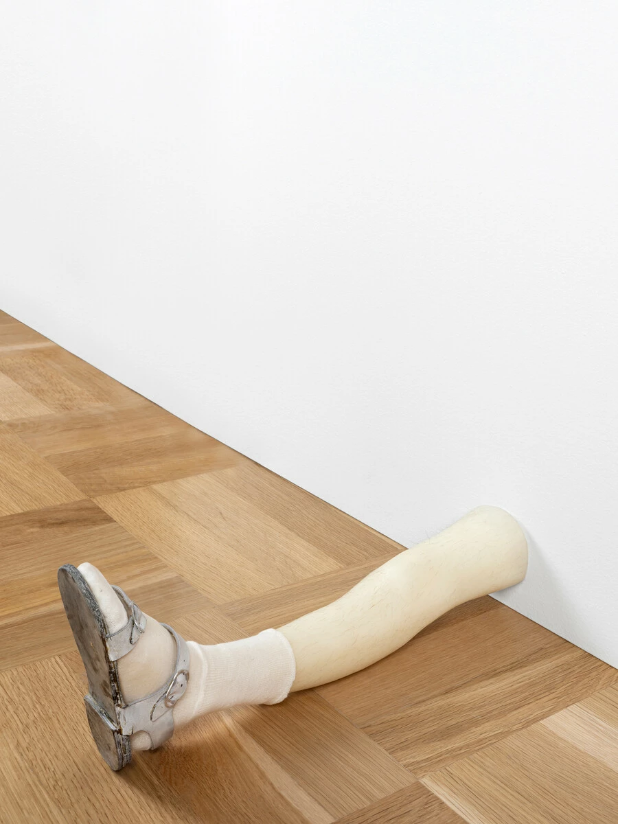A prosthetic leg wearing a sandal rests on the floor against a white wall.