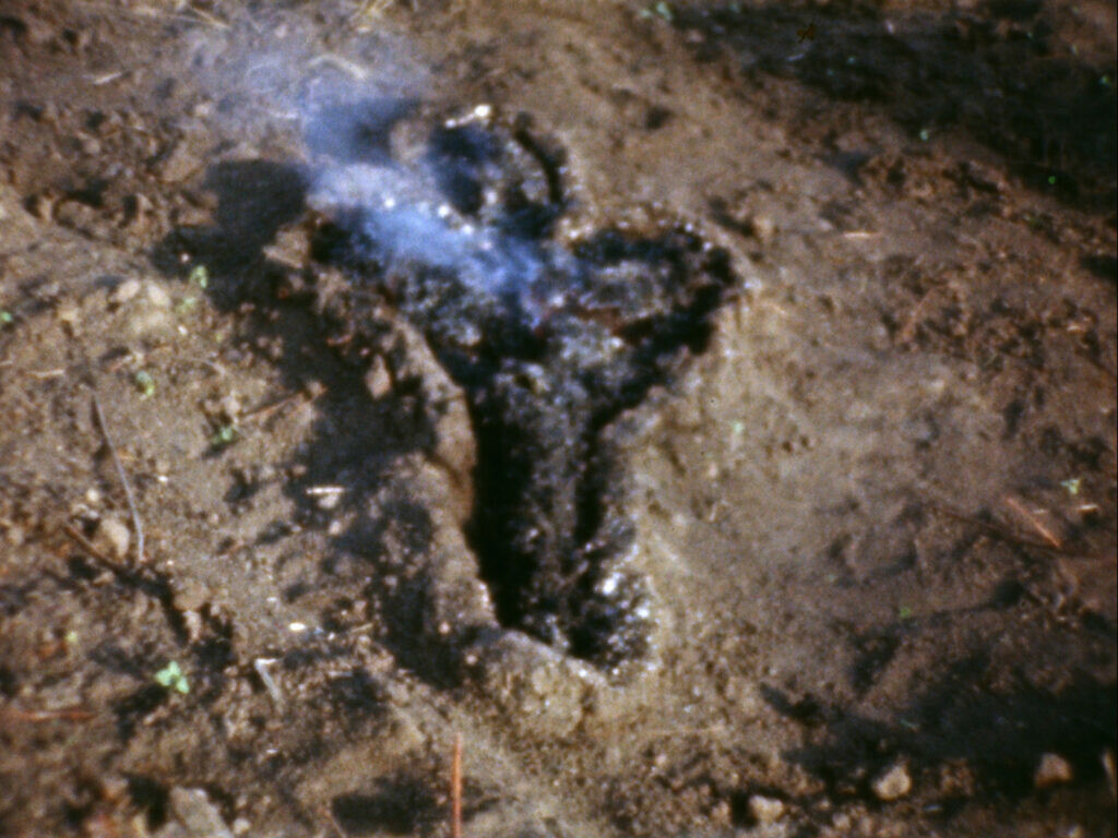 Smoke rises from a dark pile formed in the outline of a human amid a landscape of dirt.