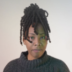 Photo of a Black person with black dreadlocks in an updo hairstyle, wearing a black turtleneck shirt. 
