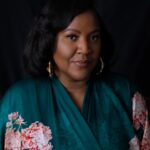 Image of a Black person with long black hair, silver earrings, and a silk flowery teal shirt. 