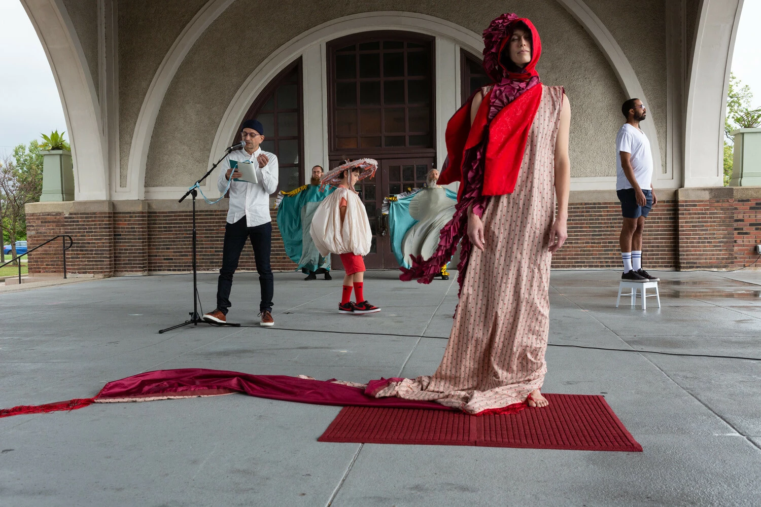 A person in a dress with a long train and red bonnet stands barefoot on a rug as a group of people in various costumes pose behind them