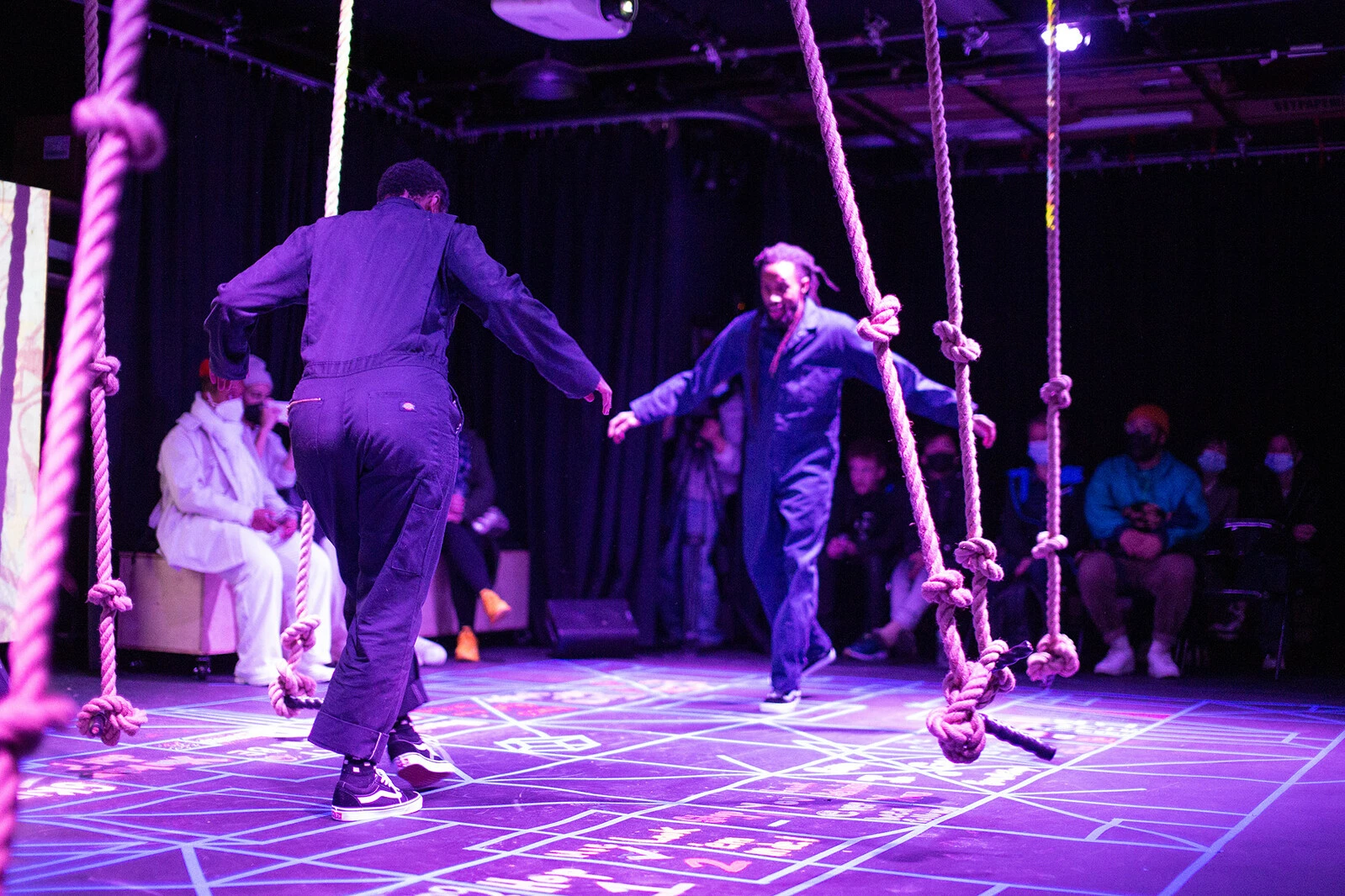 Two people facing each other among a purple-lit space full of thick, knotted ropes hanging down.