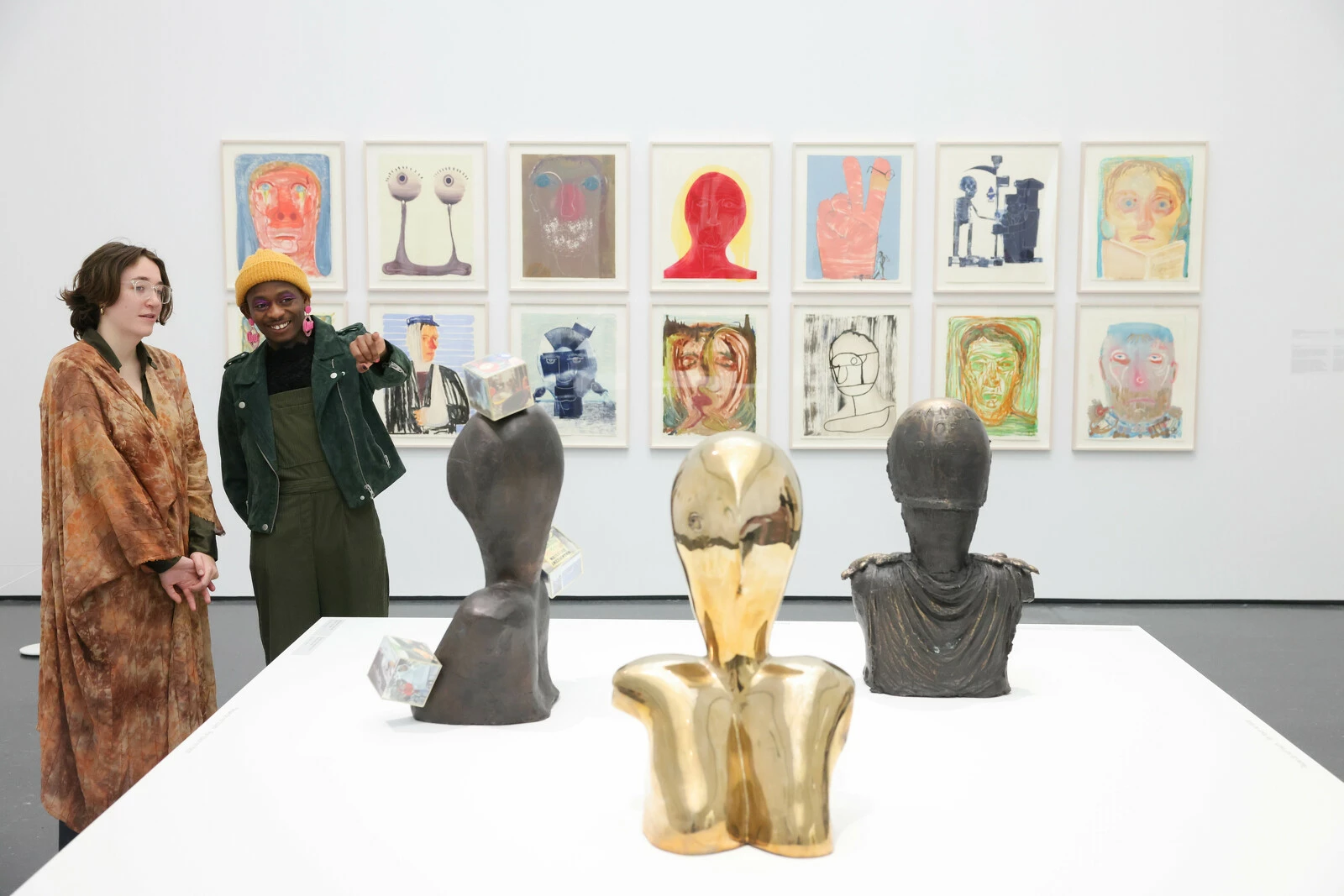 Two people look at a set of three sculptures in a gallery space with a series of drawings on the wall behind them.