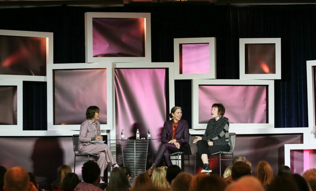 Three women sit on a stage surrounded by white-framed TVs displaying pink fabric