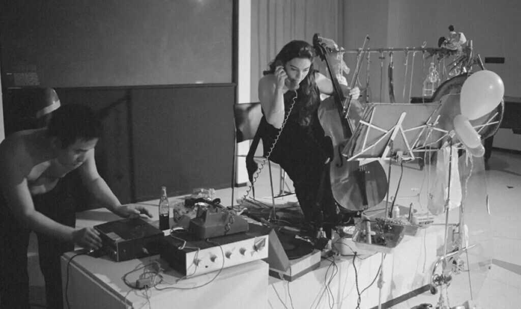 Archival photo of two people interacting with various sound-making objects, including balloons, a cello, and a record player, while one person holds a telephone receiver to their ear.