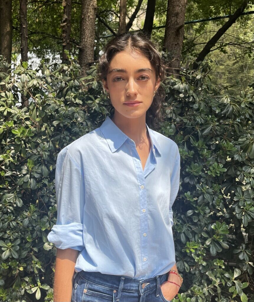 A portrait of a person dressed in a blue collared shirt and jeans with their hair pulled back in a forest setting