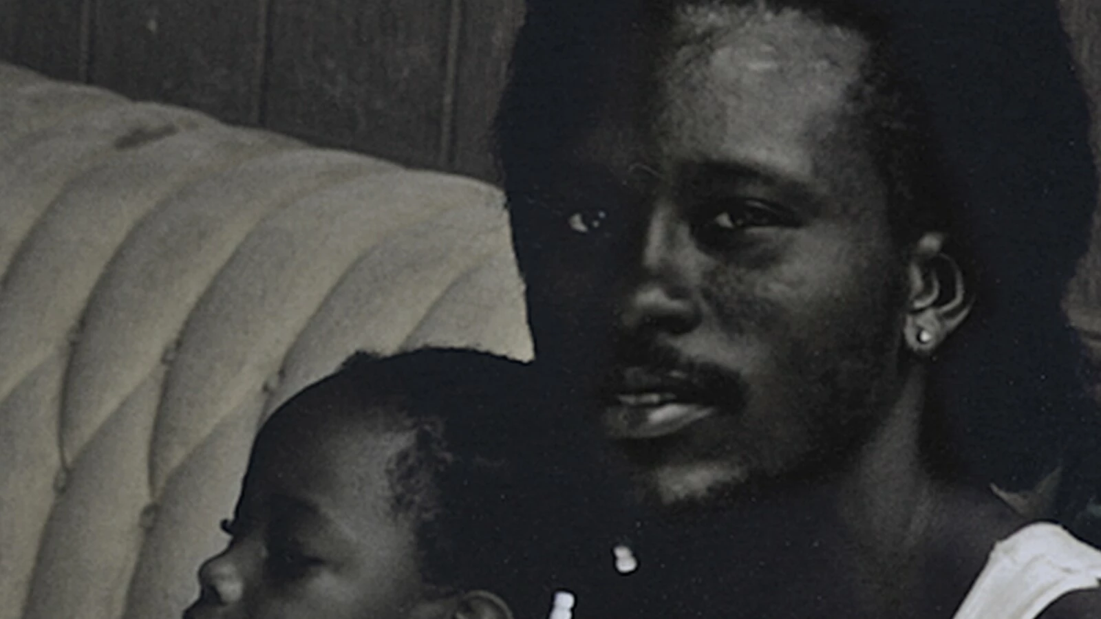 Black and white grainy image of a man with dark skin holding a small child on a couch