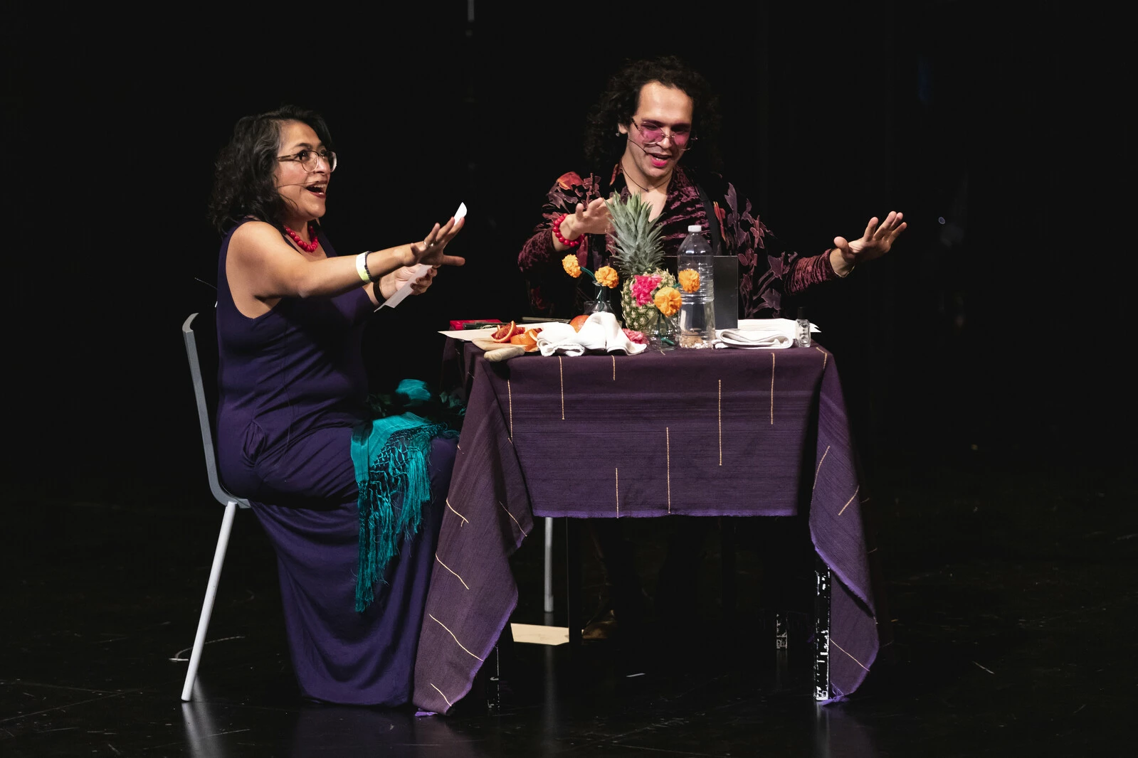 Two actors sitting at a table on a black stage