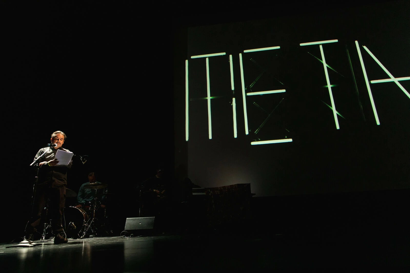 A person stands at a microphone with a band barely visible in the background. The word Meta is displayed on a large screen