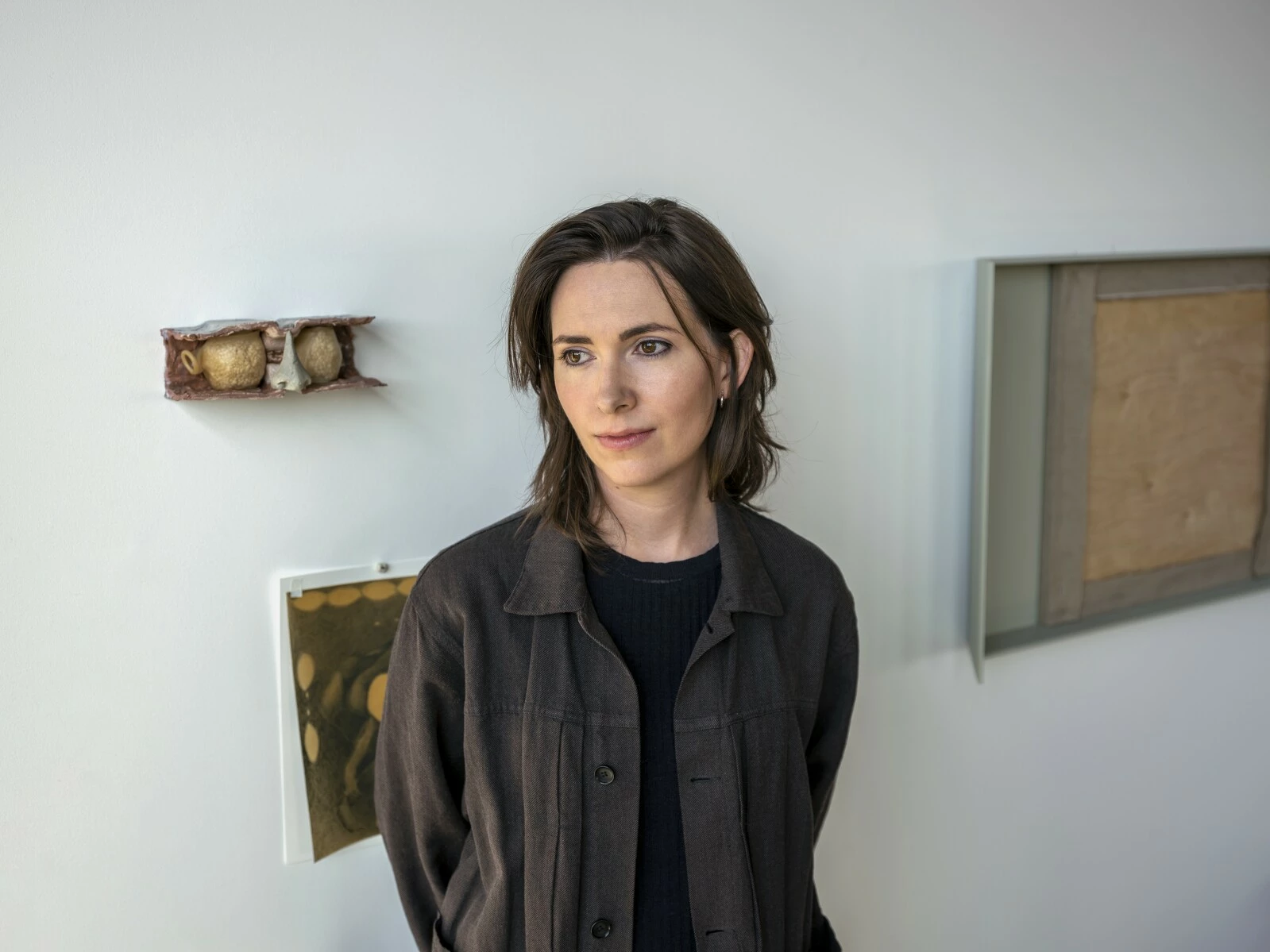 A person with medium length hair stands in front of a white wall hung with three artworks