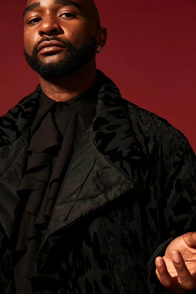 Portrait of a man with dark skin dressed in a black coat against a red wall with his left arm gesturing towards the camera