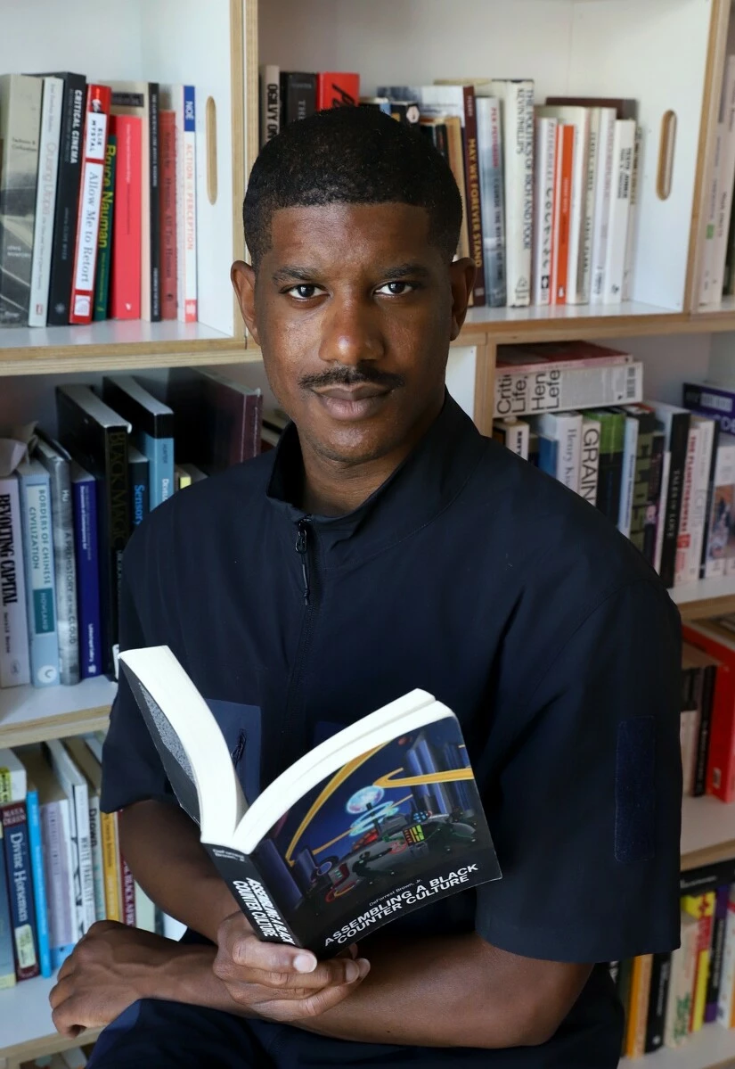 A dark-skinned man holding a book in front of a wall of books