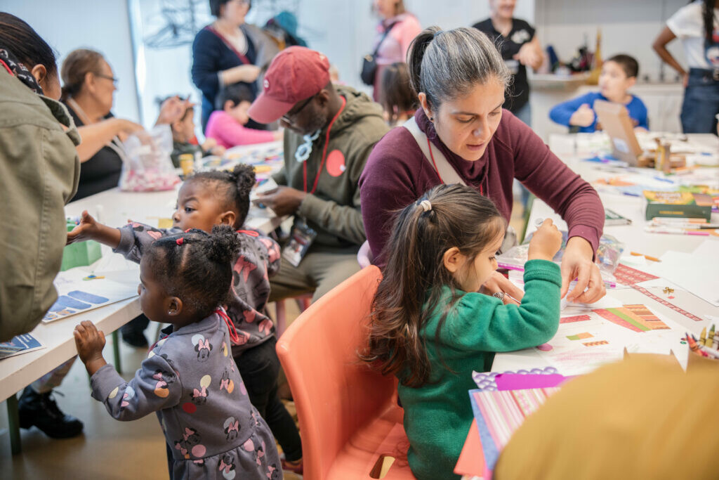 Niños y adultos se sientan en varias mesas participando en actividades artísticas