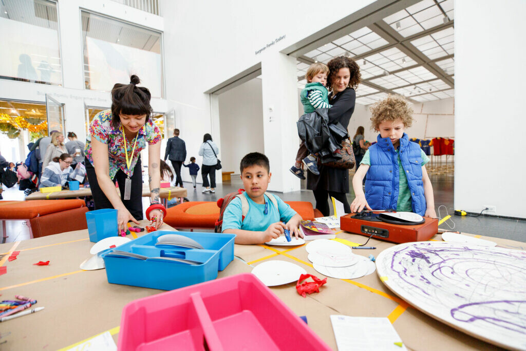 Kids and adults gather at a table filled with artmaking supplies