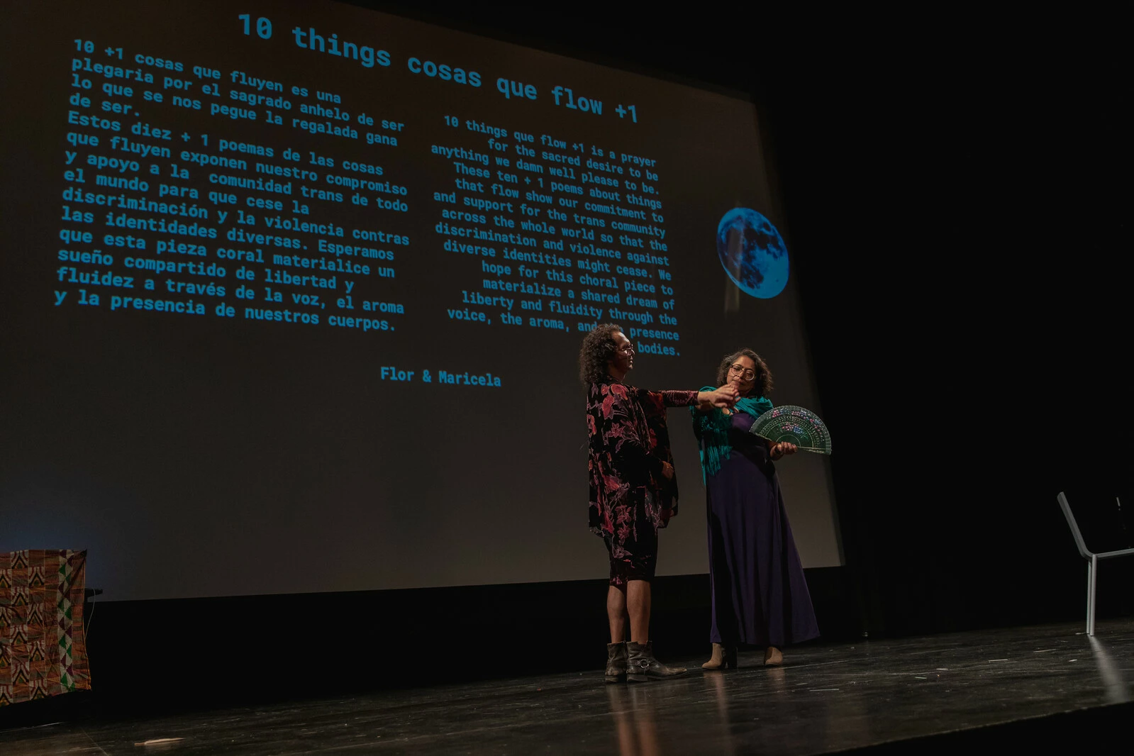 Two people interact with each other on stage in front of a screen displaying a lot of text