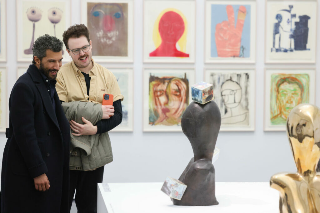 Two people look at a set of two sculptures in a gallery space with a series of drawings on the wall behind them.