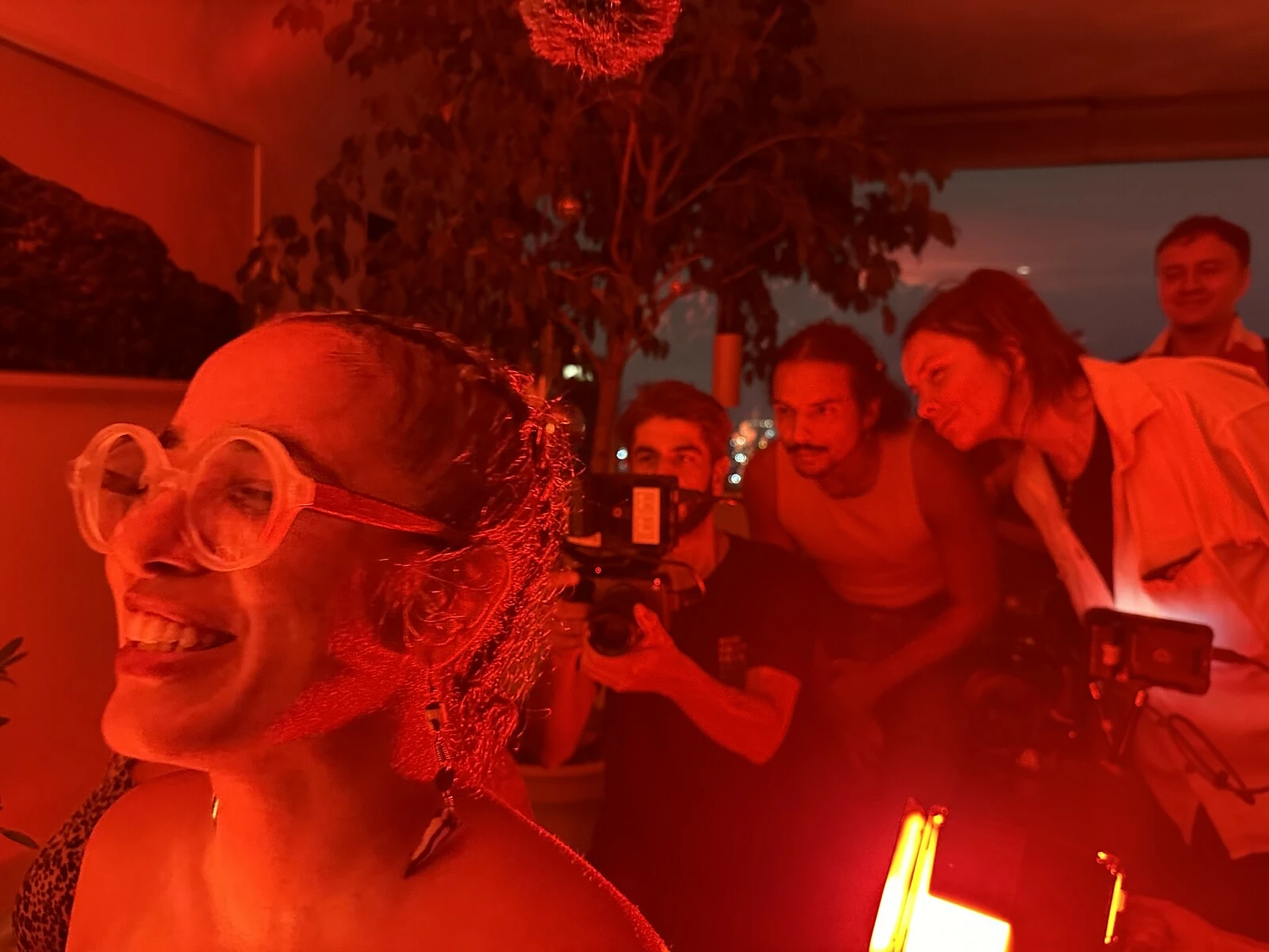 A red-tinged image of a smiling person wearing glasses in the foreground with a group of four people behind them looking at the small screen of a video camera.