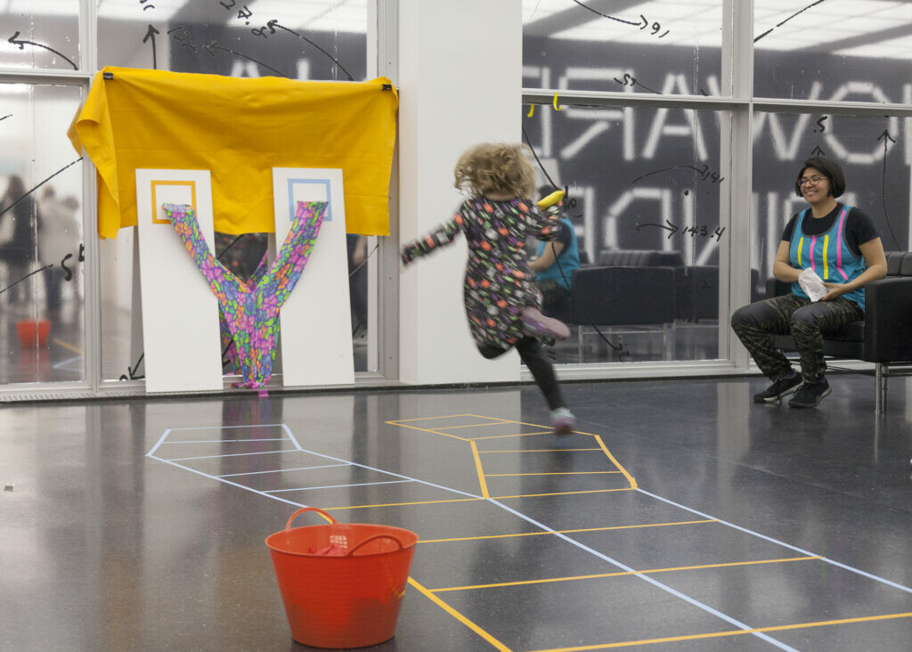 A child in midair aims a beanbag at a makeshift goal as an adult watches on.