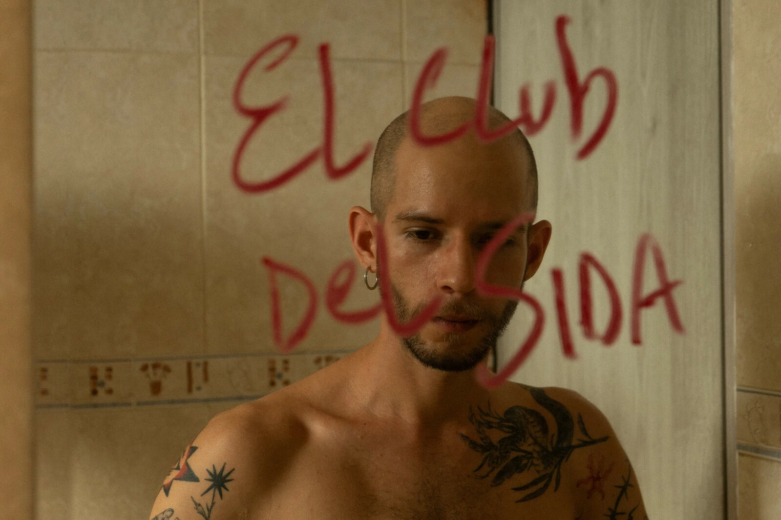 A balding, shirtless person reflected in a mirror that has on it El Club Del SIDA written in red