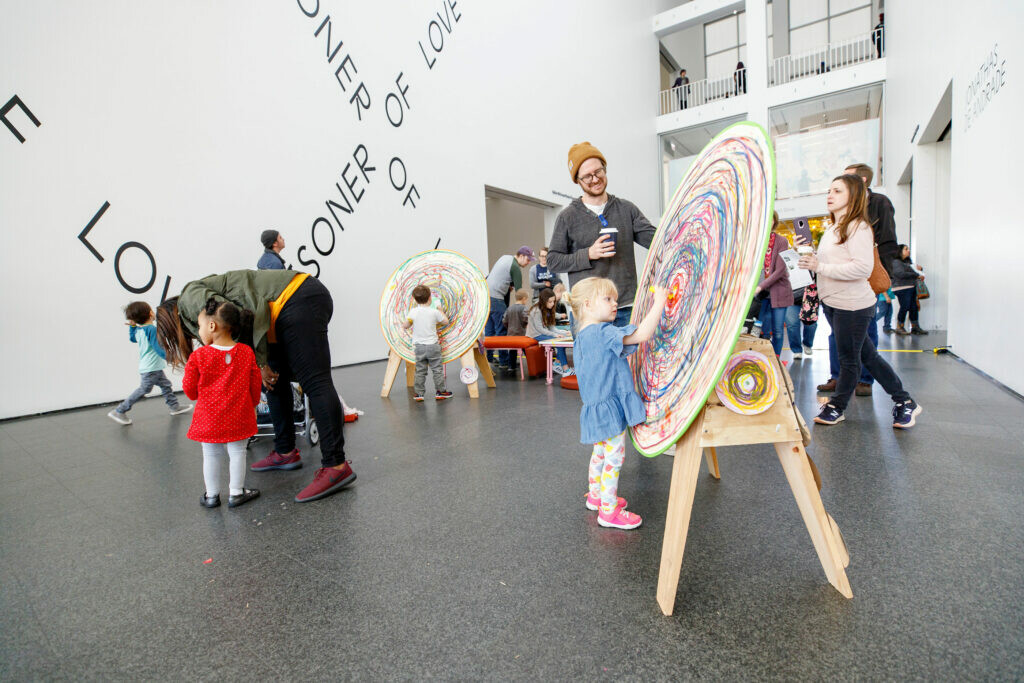 Kids and adults gather in an open, two-story space, many around easels will round canvases