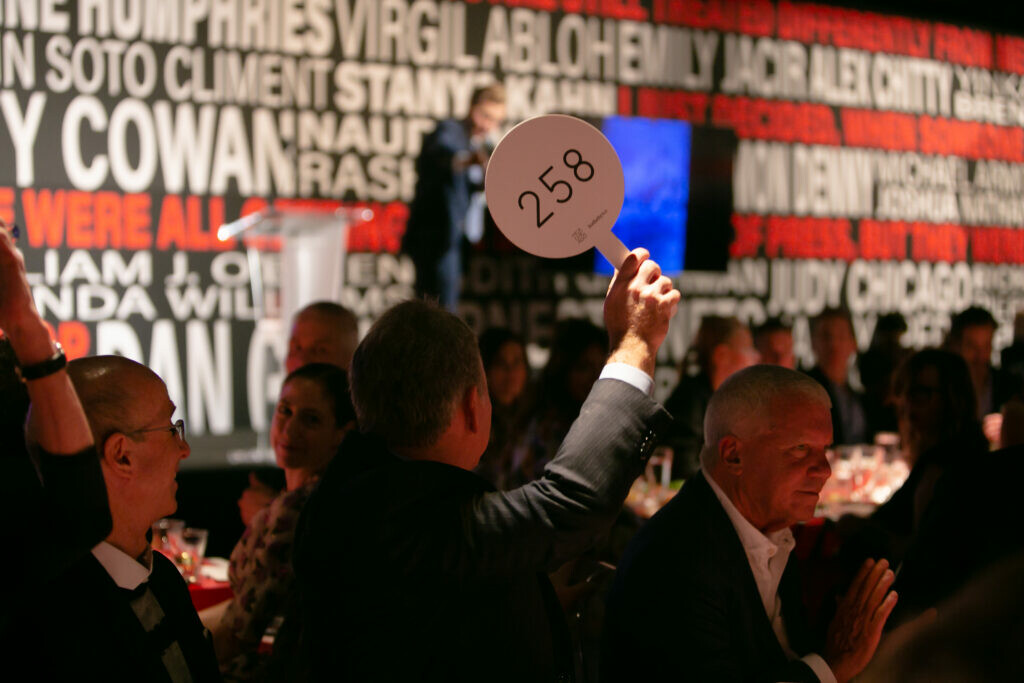 View of a seated crowd in front of a stage with one person holding up an auction paddle numbered 258