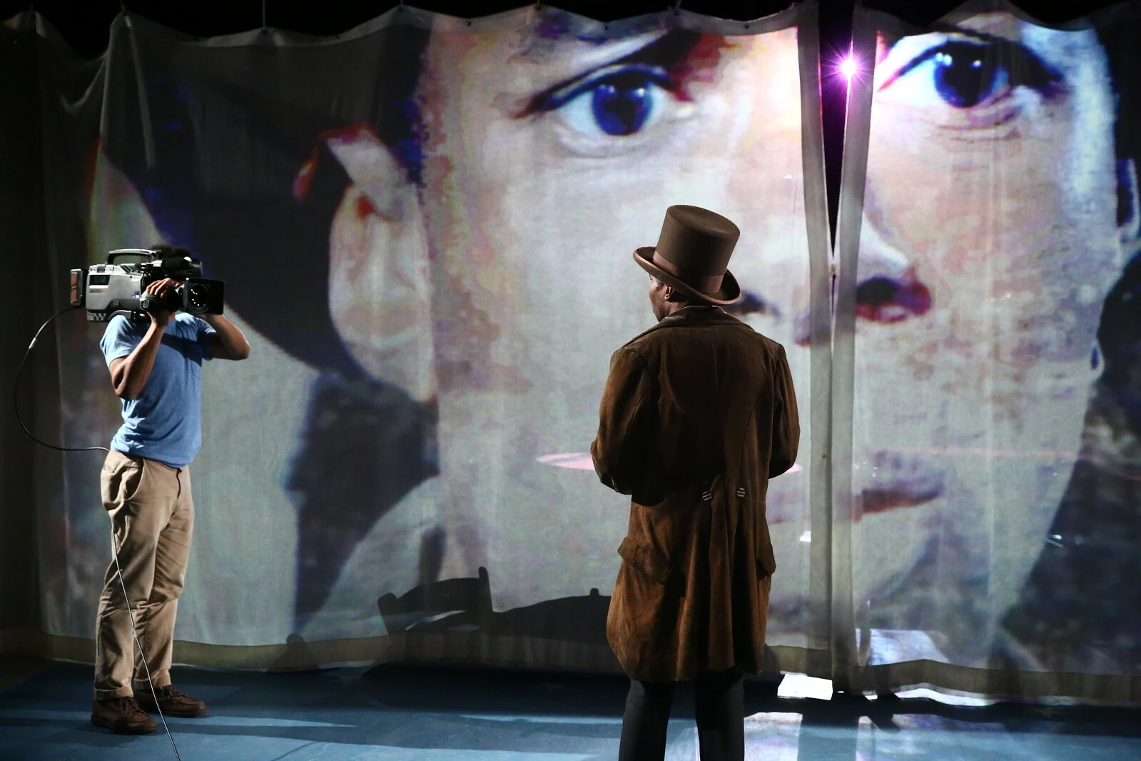 A person turned away from the camera in a top hat looks towards another person holding a video camera and a large screen displaying a man's head