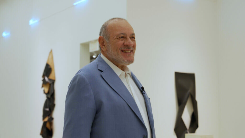 A light-skinned man in a grey suit in a gallery with art hanging on the walls behind him.