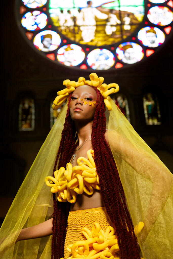 A female presenting person with light brown skin dressed in yellow with a yellow veil below a rosette style window.